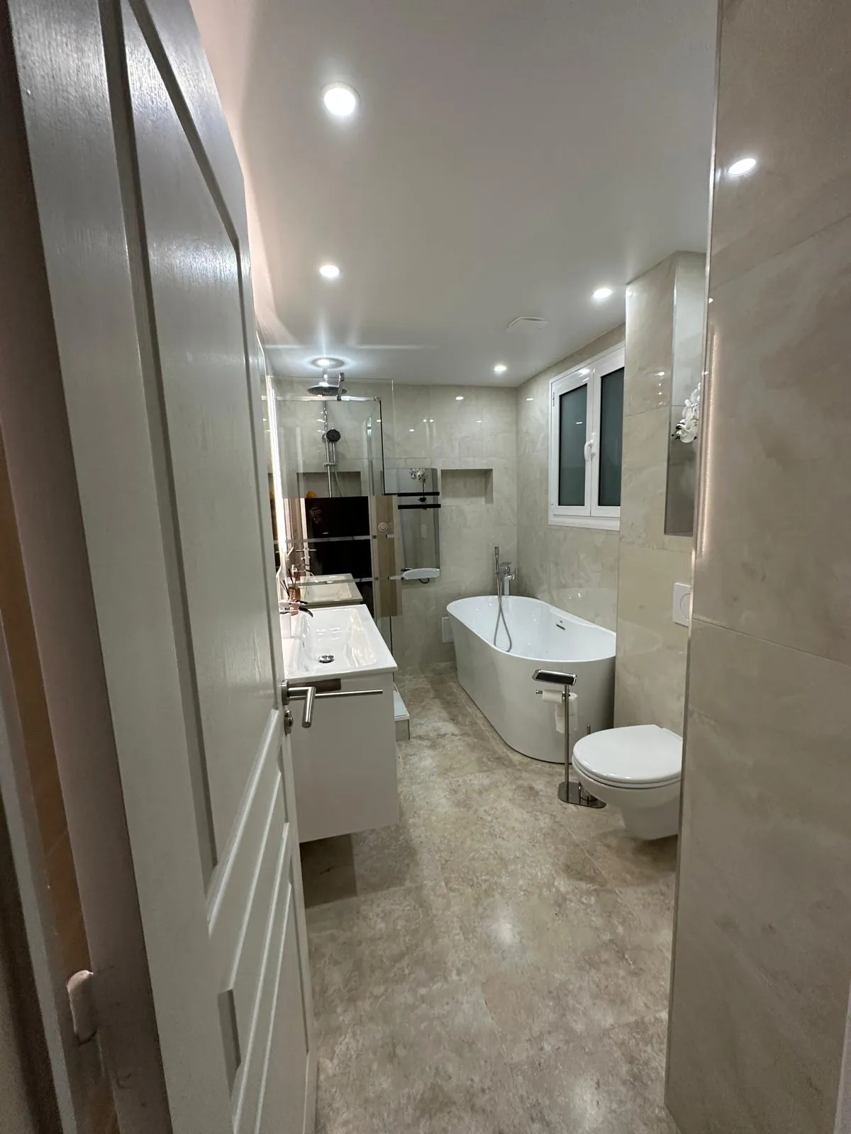 Salle de bains rénovée avec une baignoire blanche, une douche vitrée et un carrelage beige.