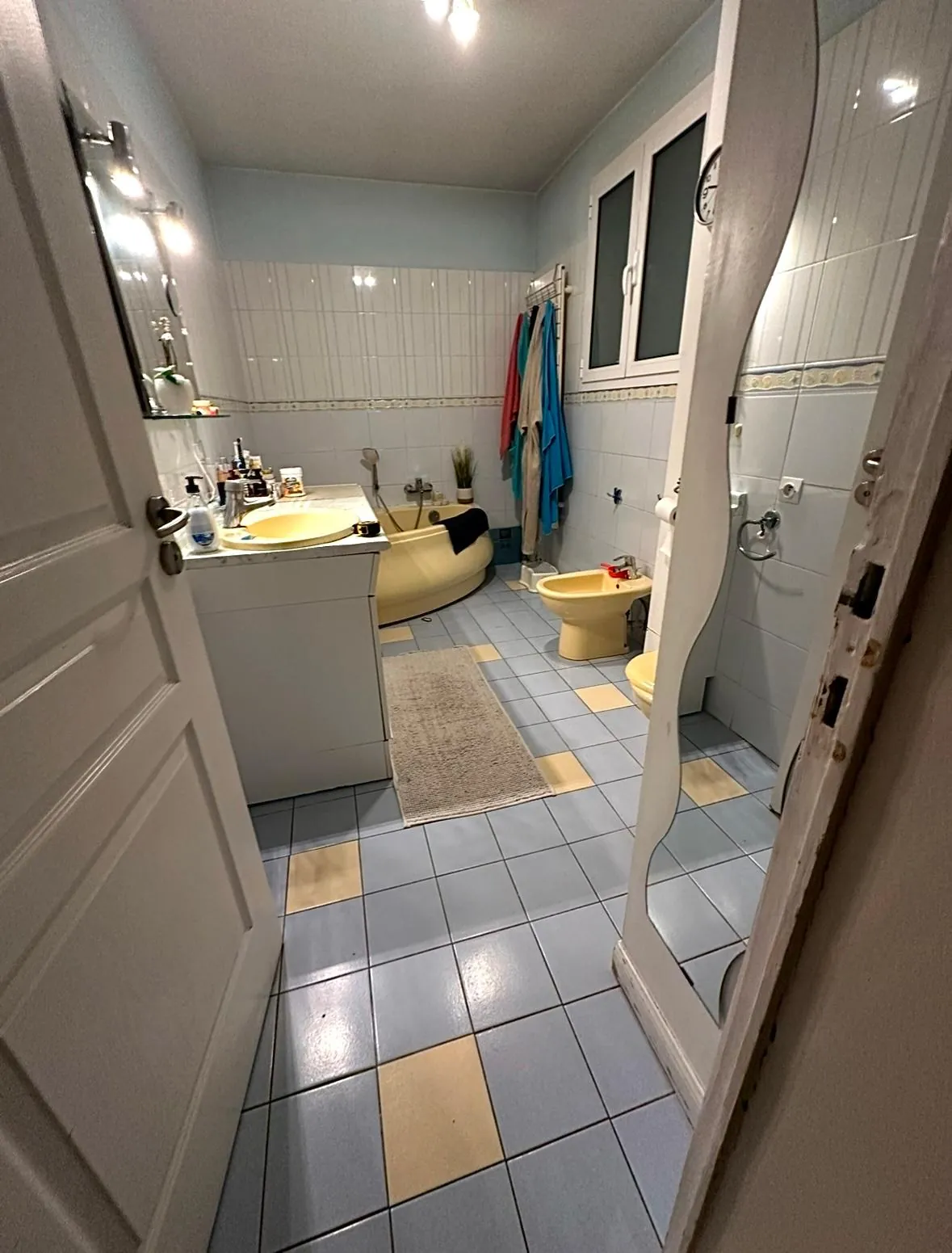 La même salle de bains avant rénovation : carrelage ancien, équipements jaunes et éclairages muraux.