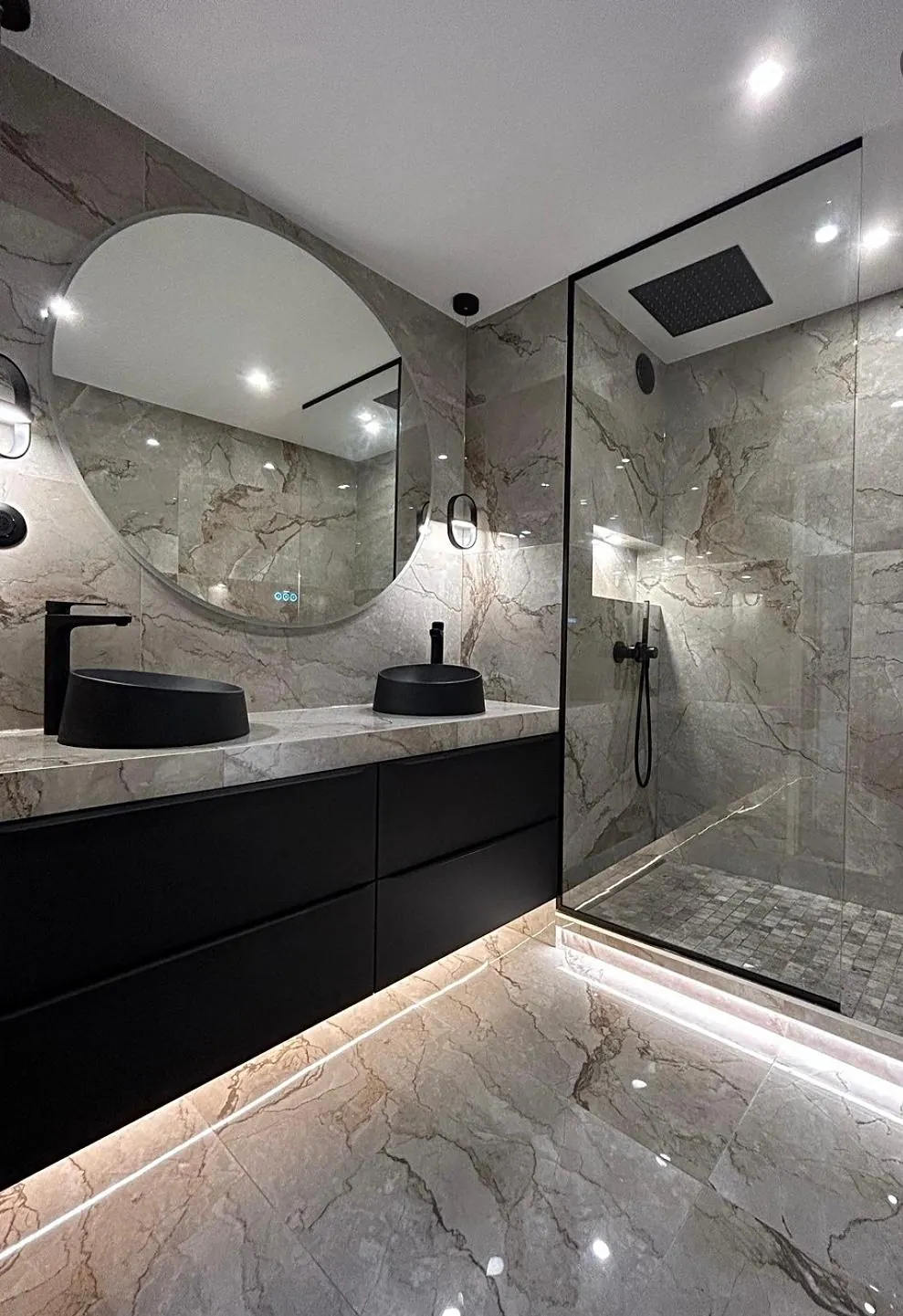 Double vasque noire, miroir rond et douche vitrée dans une salle de bains aux revêtements veinés.